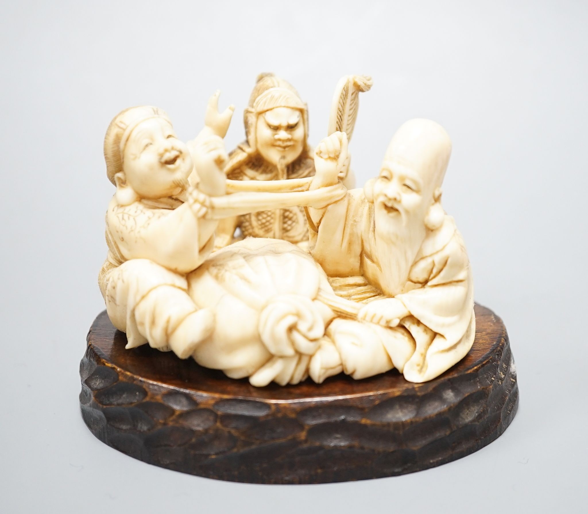 A Japanese ivory okimono of the gods Daikoku, Bishamonten and Fukurokuju, Meiji period, signed to a red lacquer tablet, 8cm wide, wood stand